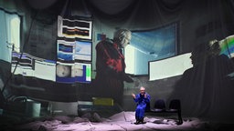 Szenenfoto von "Polaris": Ein Mann im blauen Overall vor einer überdimensionierten Projektion von Personen vorzahlreichen Monitoren im Inneren einer Forschungseinrichtung.