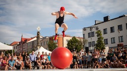 Roman Škadra springt, umgeben von Zuschauenden inmitten eine Platzes, auf einem großen roten Gymnastikball.