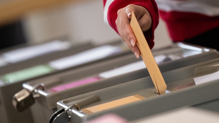 Symbolbild. Eine Hand wirft einen Wahlzettel in eine Wahlurne.