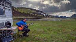 Ein Mann sitzt vor einem Camper in der Natur und spielt Gitarre.