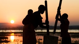 In der untergehenden Sonne steht eine Familie am Rande eines Flusses im Gegenlicht. 