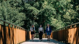 Drei junge Frauen spazieren am 19.8.2025 über eine Brücke in Mühlheim. Im Hintergrund sind sattgrüne Bäume.
