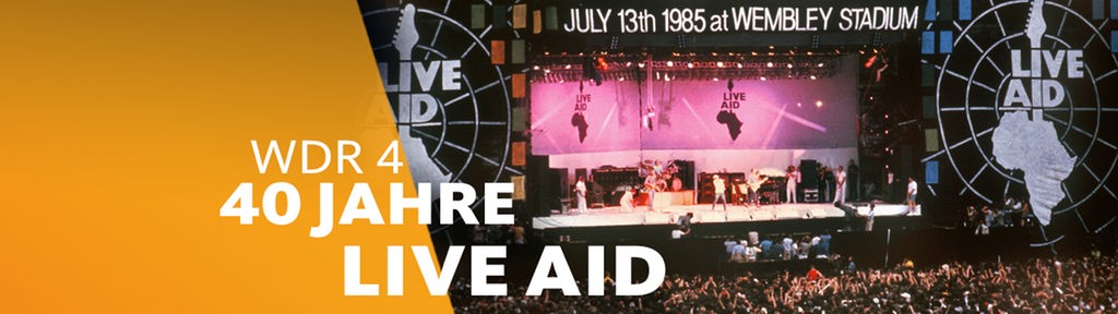  Live-Aid-Festival 1985 Wembley: Blick auf die Bühne, Menschenmenge im Vordergrund, rechts groß das Logo der Aktion