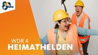 Katharina te Uhle und Bastian Bender posieren mit orangefarbener Warnweste, gelbem Bauarbeiter-Helm und Spaten vor der Kamera