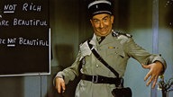 Der französische Schauspieler und Komiker Louis de Funès steht als Gendarm verkleidet vor einer beschriebenen Schultafel
