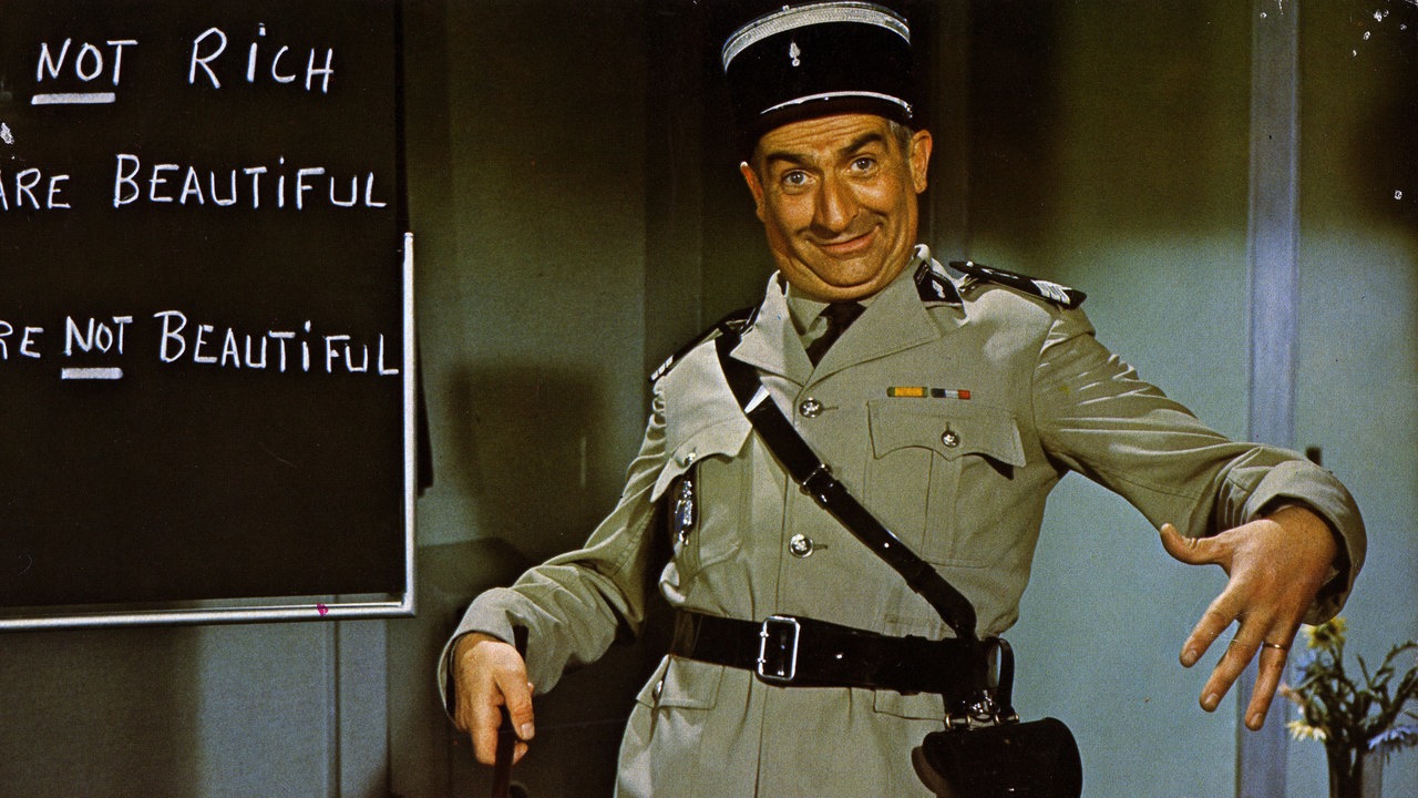 Der französische Schauspieler und Komiker Louis de Funès steht als Gendarm verkleidet vor einer beschriebenen Schultafel