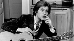Joe Dassin in den 60er Jahren mit nachdenklichem Blick und Gitarre auf dem Schoß