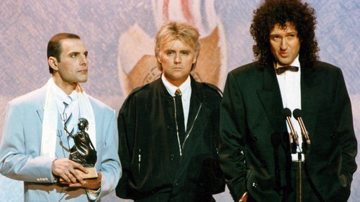 Queen 1990 bei den Brit Awards: Freddie Mercury, Roger Taylor und Brian May stehen mit ernster Miene auf der Bühne