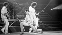 Queen live 1986 in Brüssel, Freddie Mercury posiert kniend mit Mikrofon zwischen John Deacon und Brian May auf der Bühne