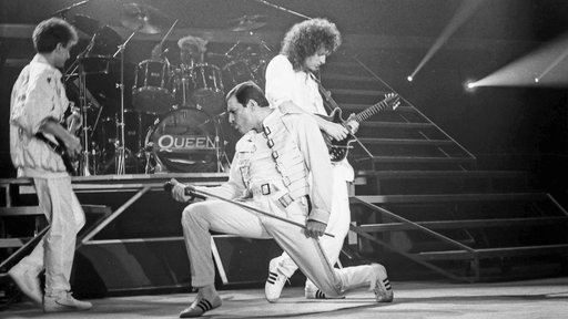 Queen live 1986 in Brüssel, Freddie Mercury posiert kniend mit Mikrofon zwischen John Deacon und Brian May auf der Bühne