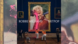 Das Cover des Albums "Britpop" zeigt ein Porträt von Robbie Williams, das in einem Museum hängt und von Aktivisten in orangenen T-Shirts mit der Aufschrift "Just Stop Pop!" mit pinker Farbe übergossen wird
