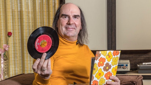 Guildo Horn auf seinem Sofa, hält eine Single und ein Aufbewahrungsalbum in den Händen