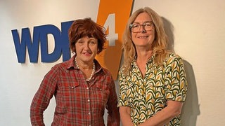 Annette Dittert und Ute Schneider vor einer weißen Wand mit WDR 4 Logo. Annette Dittert trägt ein rotes, kariertes Hemd, Ute Schneider eine grün-gelb gemusterte Bluse. 