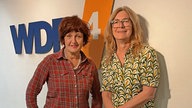Annette Dittert und Ute Schneider vor einer weißen Wand mit WDR 4 Logo. Annette Dittert trägt ein rotes, kariertes Hemd, Ute Schneider eine grün-gelb gemusterte Bluse. 