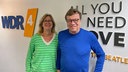 Bela Rethy und Ute Schneider im WDR 4 Studio. Bela Rethy trägt einen leuchtend-blauen Pullover, Ute Schneider einen grün-gestreiften. Die weiße Wand hinter ihnen hat einen "WDR 4"- und einen "All you need is love"- Schriftzug. 
