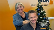 Florian Schroeder und Heike Knispel im WDR 4 Studio. Florian Schroeder sitzt am Mikrofon, Heike Knispel steht hinter ihm, die Arme auf der Stuhllehne abgestützt und verschränkt. Beide lächeln in die Kamera. Hinter ihnen steht ein Weihnachtsbaum. 