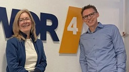Frank Busemann mit Ute Schneider bei WDR 4: Sie lehnen sich lässig an die Wand in einem weißen Flur mit dem Schriftzug "Wir sind Heimat. WDR 4" an der Wand. Beide lächeln freundlich in die Kamera.