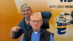 Pfarrer Franz Meurer und Heike Knispel im WDR 4-Studio. Franz Meurer sitzt am Mikrofon, Heike Knispel steht hinter ihm, einen Arm auf die Stuhllehne gelegt, mit dem anderen zeigt sie auf ihn. 