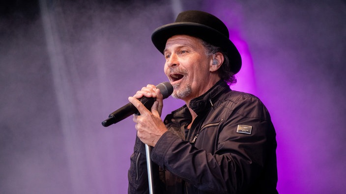 Peter Brings auf der Bühne. Er singt gerade ins Mikrofon, welches er mit beiden Händen festhält. Er trägt eine schwarze Jacke und einen schwarzen Hut. Die Bühnenbeleuchtung im Hintergrund ist lila.