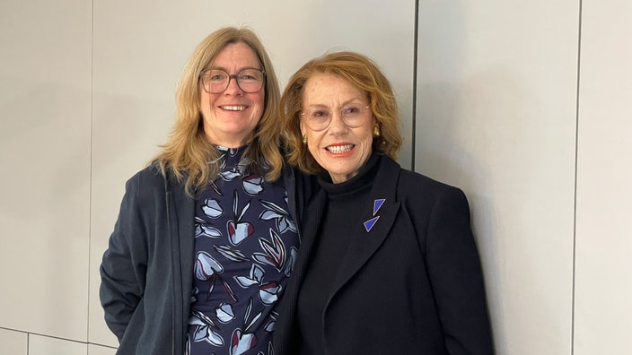 Ein Foto von Ute Schneider und Gaby Dohm. Die stehen nebeneinander vor einer weißen Wand und lächeln in die Kamera.