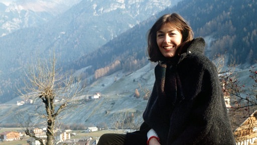 Thekla Carola Wied sitzt auf einem Stein, im Hintergrund sieht man ein Dorf in den Bergen, es ist Winter oder Herbst