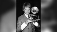 Thomas Fritsch lächelt in die Kamera und präsentiert eine Grammophon-Spieluhr