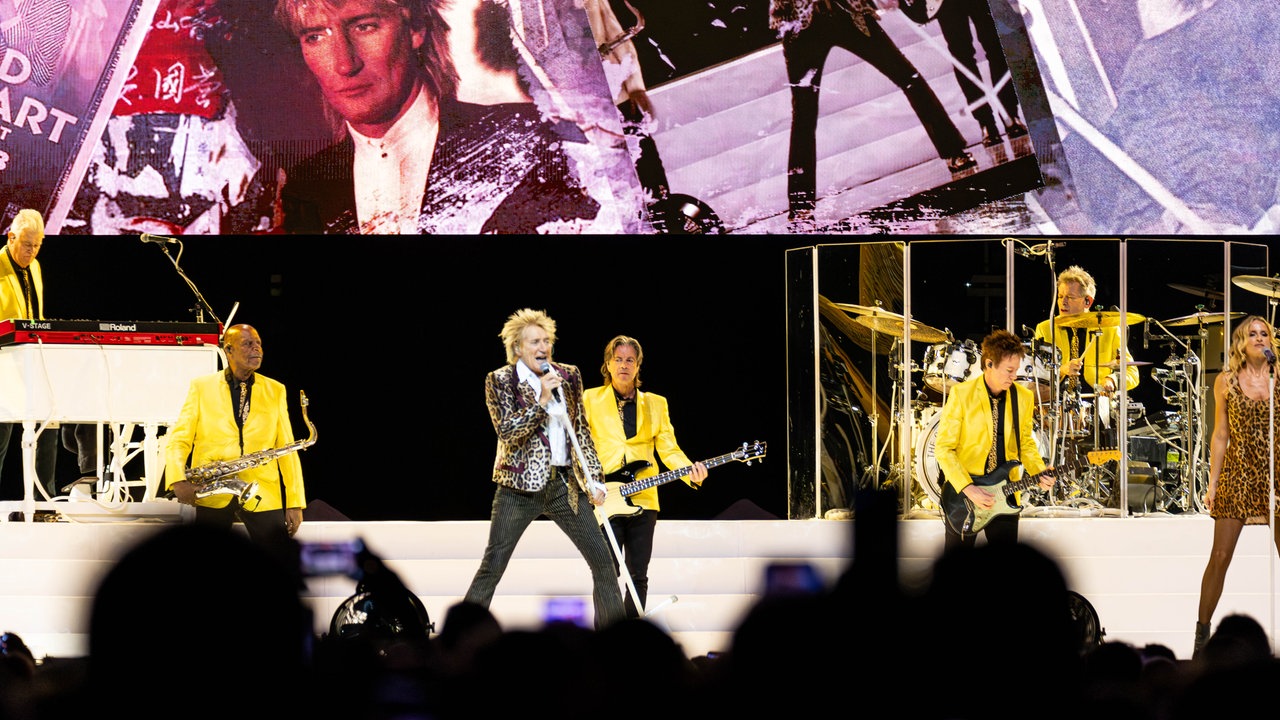 Rod Stewart singt auf einer Bühne, neben ihm spielt seine Band