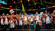 Lachende Köln Arena 2026: Junge Mitglieder eines Tanzcorps stehen in orange-weißer Uniform auf der Bühne und trommeln