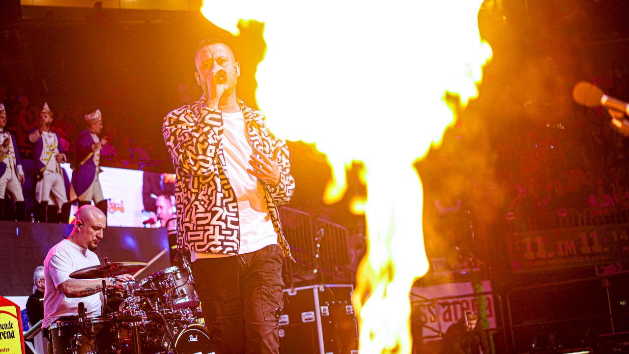 Lachende Köln Arena 2026: Sänger Mo-Torres steht mit Mikrofon auf der Bühne. Im Vordergrund ist ein Feuer von der Pyroshow der Bühne zu sehen