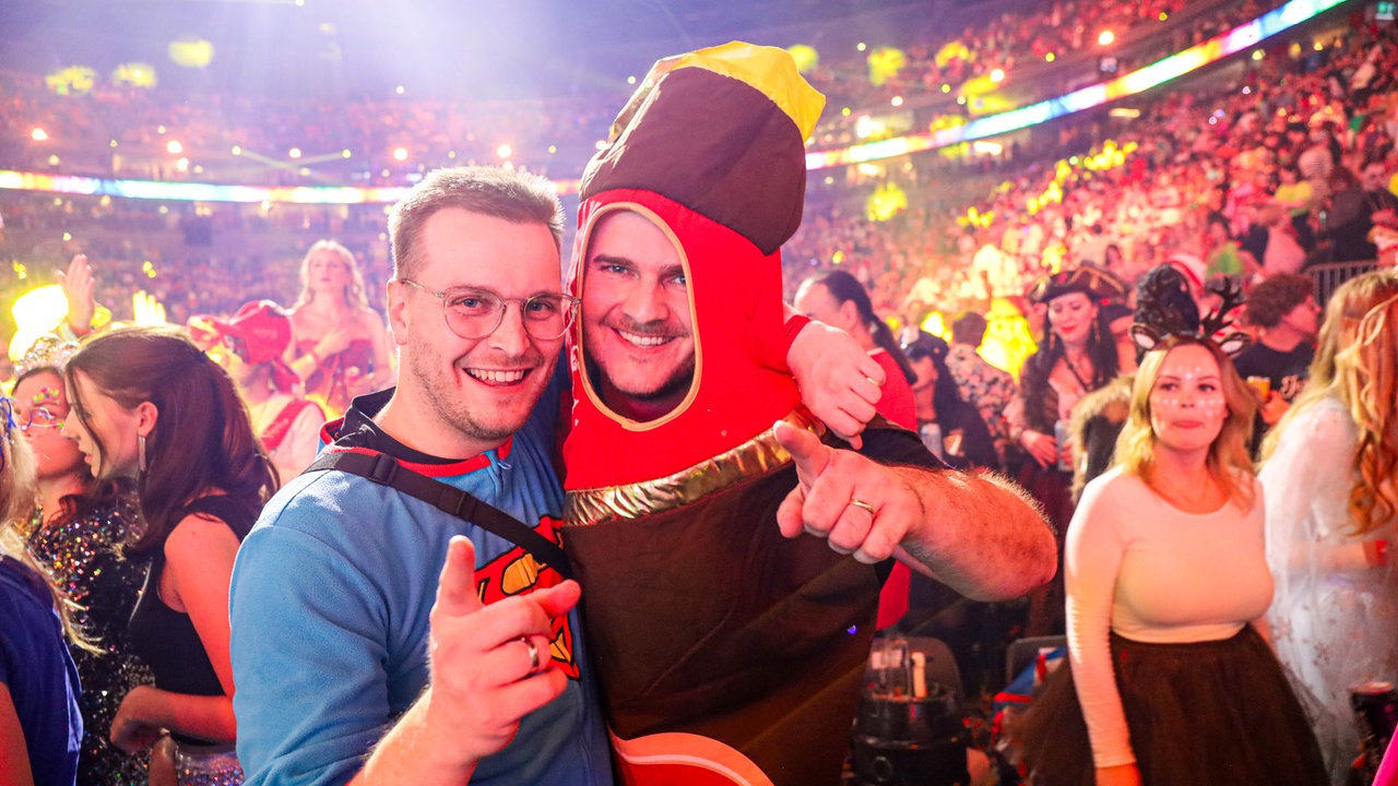 Lachende Köln Arena 2026: Zwei verkleidete Jecken. Ein Karnevalist hat sich als Superman verkleidet, sein Kollege kommt als Flasche Bier in die Arena.