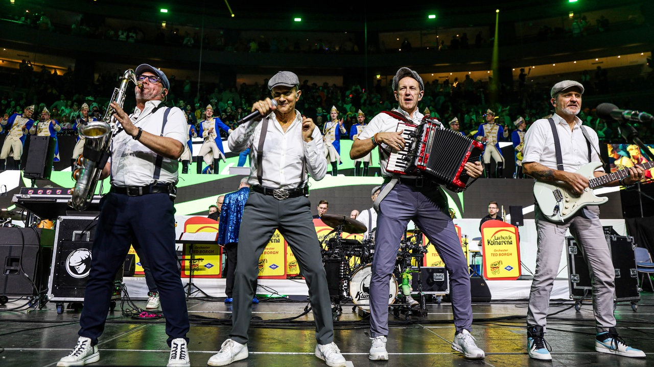 Lachende Köln Arena 2026: Vier Musiker der Band Klingelköpp stehen mit ihren Instrumenten in einem weißen Hemd und Schiebermütze auf der Bühne