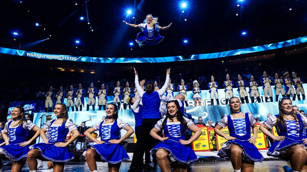 Lachende Köln Arena 2026: Ein blau-weißer Tanzcorps steht auf der Bühne. Fünf Mädchen knien, mehrere Männer werfen eine Tänzerin in die Höhe
