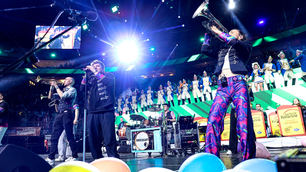 Lachende Köln Arena 2026: Auftritt der Band Druckluft. Zwischen Trompetern steht der Sänger auf der Bühne. Im Vordergrund sind bunte Luftballons