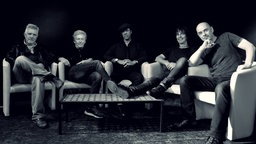Schwarz-weiß Foto der Manfred Mann's Earth Band, auf Sesseln um einen Couchtisch herum sitzend.