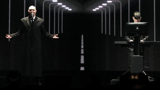 Die Pet Shop Boys auf der Bühne: Das Bühnenbild ist dunkel gehalten, beide sind schwarz gekleidet. 