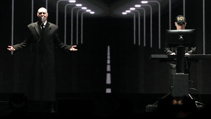 Die Pet Shop Boys auf der Bühne: Das Bühnenbild ist dunkel gehalten, beide sind schwarz gekleidet. 