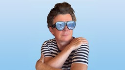 Rick Astley in gestreiftem T-Shirt mit Sonnenbrille vor blauem Hintergrund