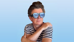 Rick Astley in gestreiftem T-Shirt mit Sonnenbrille vor blauem Hintergrund