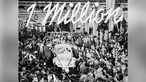 Im VW-Werk in Wolfsburg wird die Fertigstellung des millionsten Käfers gefeiert. Hunderte Menschen stehen um das Fahrzeug herum, im Hintergrund ist ein großer Schriftzug '1 Million' zu sehen