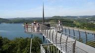 Eine runde Aussichtsplattform mit Ausblick über den Biggesee. Es ist sonnig, man sieht mehrere Personen auf der Plattform. 