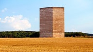 Ein Stoppelfeld, die Bruder-Klaus-Feldkapelle in Mechernich mittig im Bild, im Hintergrund sind Bäume und ein blauer Himmel zu sehen. Die Kapelle ist keine traditionelle Kapelle, sondern eher minimalistisch gehalten, sie ist fensterlos und eckig gebaut. 