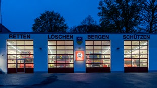 Außenansicht des Gerätehauses der Freiwilligen Feuerwehr Breckerfeld in der Dämmerung. Über den Eingängen stehen die Worte "retten, löschen, bergen, schützen". Die Löschfahrzeuge stehen im beleuchteten Gerätehaus. 