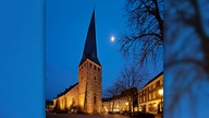 Der schiefe Turm der Georgskirche in Hattingen in der Dämmerung. Die Kirche und umliegenden Gebäude sind warm beleuchtet, am Himmel sieht man den Mond.