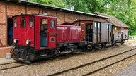 Eine rote Diesellok steht auf den Schienen vor einem Güterschuppen. 