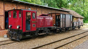 Eine rote Diesellok steht auf den Schienen vor einem Güterschuppen. 