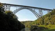 Die Müngstener Brücke zwischen Remscheid und Solingen: Eine stählerne Bogenbrücke vor blauem Himmel. Darunter fließt die Wupper, rechts und links sieht man Bäume und Grünflächen.