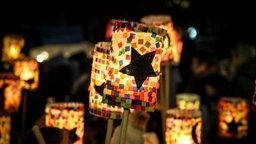 Leuchtende, selbstgebastelte Kinderlaternen am Abend: Im Fokus steht eine zylinderförmige Laterne, die mit einem großen Stern in der Mitte und bunten Papierschnipseln drumrum beklebt ist. Im Hintergrund sieht man verschwommen weitere ähnliche Laternen. 