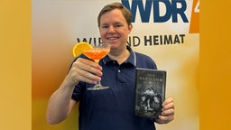 Mike Altwicker im WDR 4-Studio mit dem Buch "Der Barmann des Ritz" von Philippe Collin in der einen und einem Cocktail in der anderen Hand