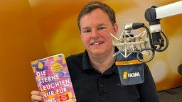 Mike Altwicker sitzt im WDR 4 Studio mit dem Buch "Die Sterne leuchten nur für uns" von Virginie Grimaldi in der einen Hand und in der anderen hält er einen Teller mit Muffins.