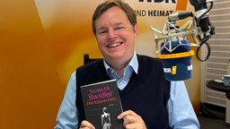 Mike Altwicker sitzt im WDR 4-Studio und hält das Buch "Herzlauschen" von Nicole und Uli Swidler in der Hand